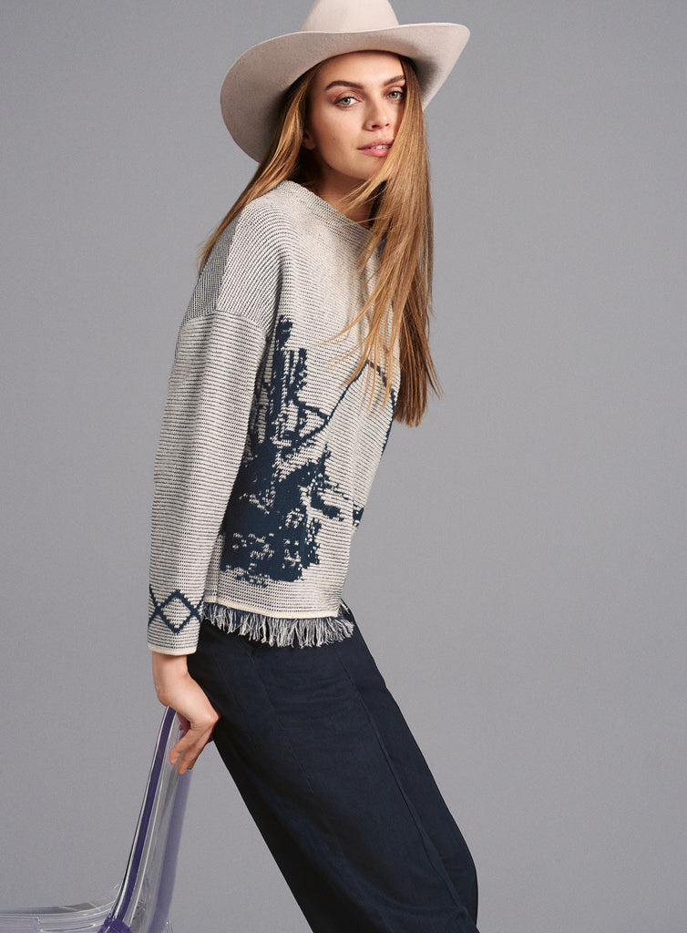 Ein weibliches Model mit langen, offenen Haaren und beigefarbenem Hut trägt einen hellgrauen Pullover mit dunkelblauem, abstraktem Muster auf der linken Seite. Der Pullover hat Fransen am unteren Saum sowie ein Rautenmuster an der Ärmelkante. Dazu trägt das Model eine dunkelblaue Hose und sitzt locker an der Seite eines transparenten Stuhls. Der Hintergrund ist einfarbig grau.
