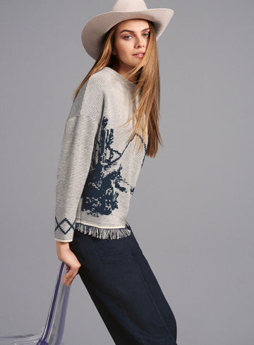 Ein weibliches Model mit langen, offenen Haaren und beigefarbenem Hut trägt einen hellgrauen Pullover mit dunkelblauem, abstraktem Muster auf der linken Seite. Der Pullover hat Fransen am unteren Saum sowie ein Rautenmuster an der Ärmelkante. Dazu trägt das Model eine dunkelblaue Hose und sitzt locker an der Seite eines transparenten Stuhls. Der Hintergrund ist einfarbig grau.