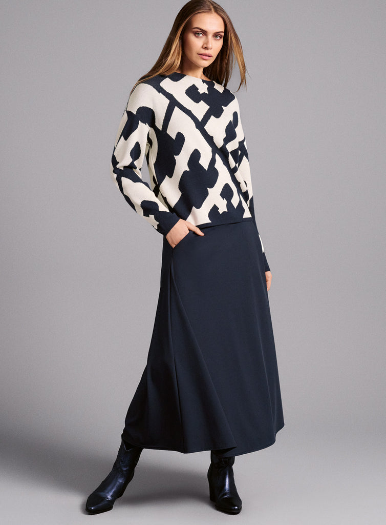 Modelansicht: Ein gestrickter Pullover mit abstraktem, kontrastreich dunkelblauem und weißem Muster. Die Grundfarbe ist Weiß, das Muster besteht aus großen, unregelmäßigen dunkelblauen Formen. Der Pullover hat einen Rundhalsausschnitt, lange Ärmel und eine lockere Passform.