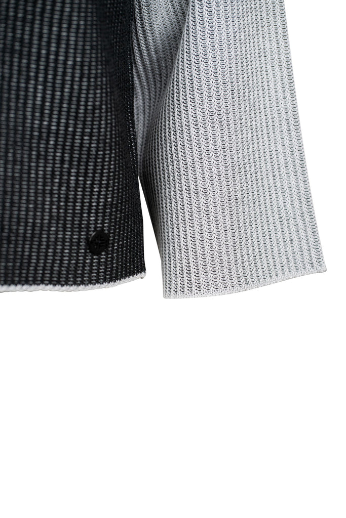 Detailansicht: Grob gestrickter Damenpullover mit asymmetrischem Farbmuster in Hellgrau und Schwarz. Die rechte Seite, einschließlich Ärmel und Schulter, ist hellgrau; die linke Seite ist überwiegend schwarz. Die Farben verlaufen diagonal über die Vorderseite. Der Pullover hat einen weiten Rundhalsausschnitt und locker fallende, gerade Ärmel.