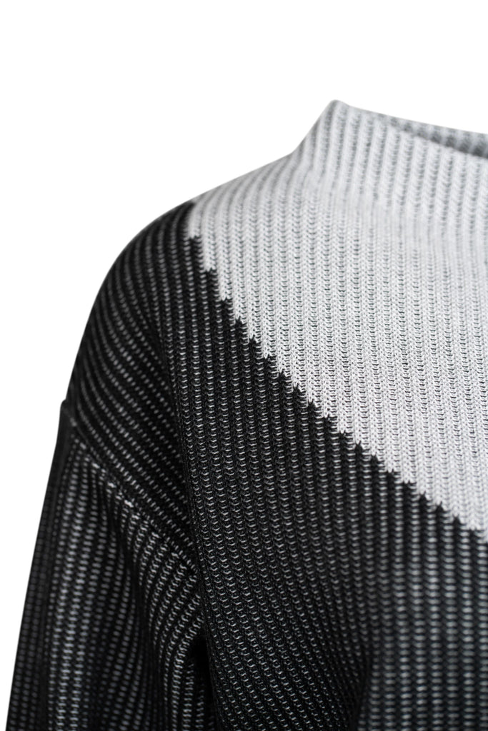 Detailansicht: Grob gestrickter Damenpullover mit asymmetrischem Farbmuster in Hellgrau und Schwarz. Die rechte Seite, einschließlich Ärmel und Schulter, ist hellgrau; die linke Seite ist überwiegend schwarz. Die Farben verlaufen diagonal über die Vorderseite. Der Pullover hat einen weiten Rundhalsausschnitt und locker fallende, gerade Ärmel.