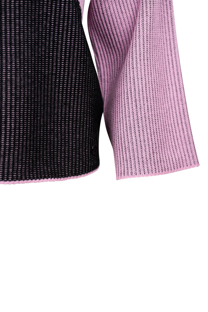Detailansicht: Ein gestrickter Pullover mit asymmetrischem Design. Die linke Seite ist überwiegend schwarz, während die rechte Seite in einem zarten Rosa gehalten ist. Die Farben treffen sich in einer geschwungenen Linie über der Brust. Der Pullover hat lange, gerade Ärmel und einen hohen Rundhalsausschnitt.