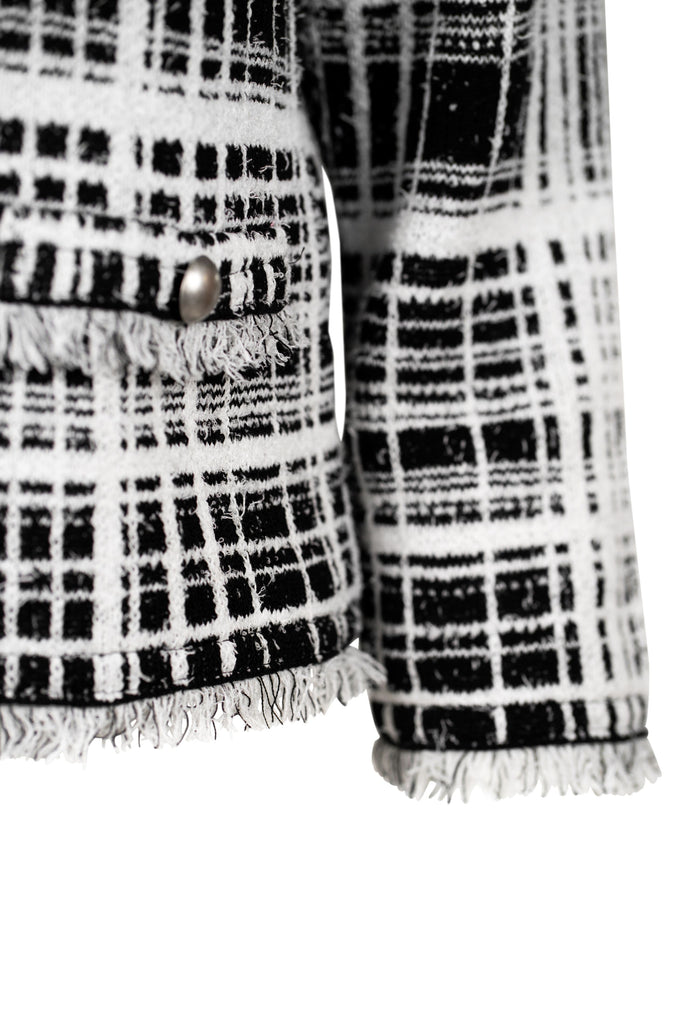 Detailansicht Arm - Klassische Bouclé-Jacke in weiß mit schwarzem Karomuster, durchgehender Knopfleiste und dekorativen Fransenabschlüssen an Kragen, Saum und Ärmeln. Zwei kleine Pattentaschen auf Hüfthöhe unterstreichen den eleganten Look im Chanel-Stil.