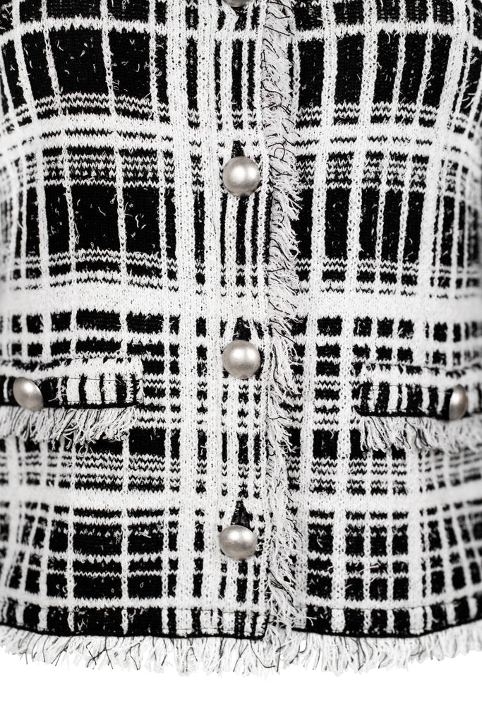Detailansicht Knopfleiste - Klassische Bouclé-Jacke in weiß mit schwarzem Karomuster, durchgehender Knopfleiste und dekorativen Fransenabschlüssen an Kragen, Saum und Ärmeln. Zwei kleine Pattentaschen auf Hüfthöhe unterstreichen den eleganten Look im Chanel-Stil.