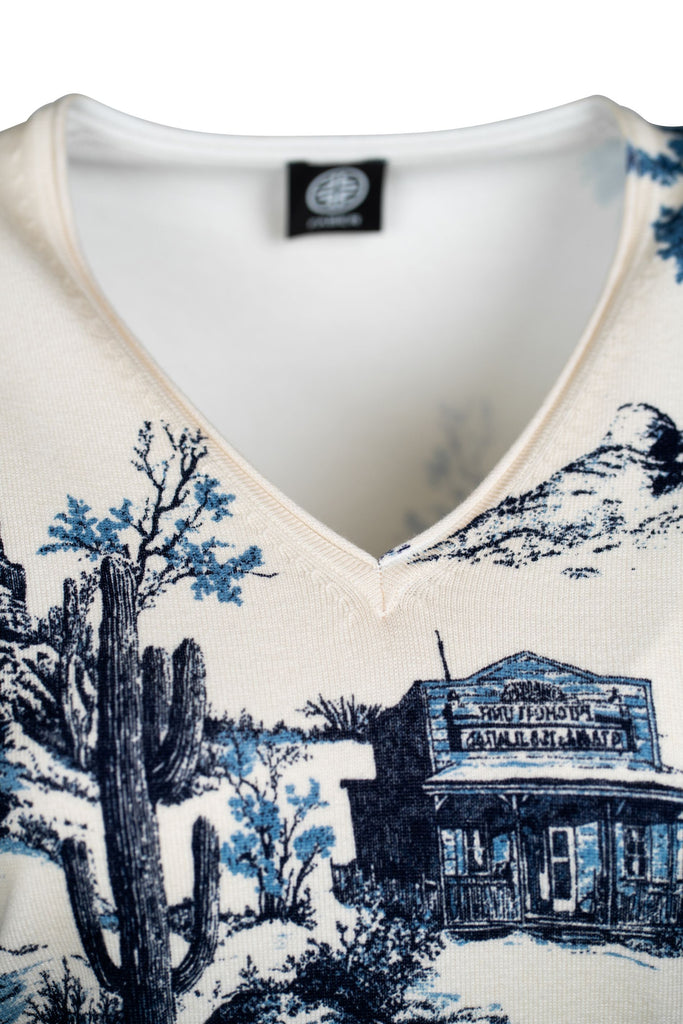 Detaillansicht des Ausschnitts des cremefarbenen Pullovers mit V-Ausschnitt und einem detaillierten, blauen Landschaftsmotiv. Das Design zeigt stilisierte Natur- und Gebäudemotive und verleiht dem Pullover einen modischen, eleganten Charakter.