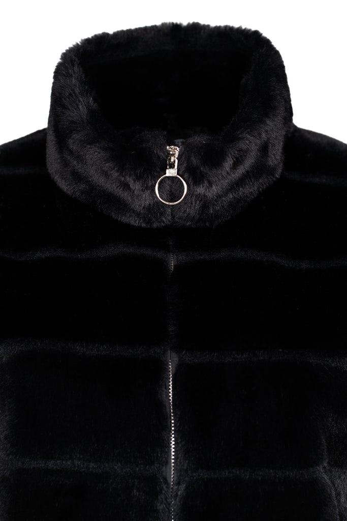 Detailansicht Schwarze, ärmellose Fellweste mit Stehkragen und durchgehendem Reißverschluss vorne. Die Weste ist in waagerechte, voluminöse Segmente unterteilt und hat ein weiches, plüschiges Obermaterial. Am Reißverschluss ist ein runder Zipper-Anhänger angebracht.